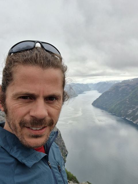 A smiling man taking a selfie above a long fjord under grey clouds.