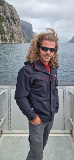 A man with long curly windswept hair and sunglasses on a ferry deck with cliffs behind.