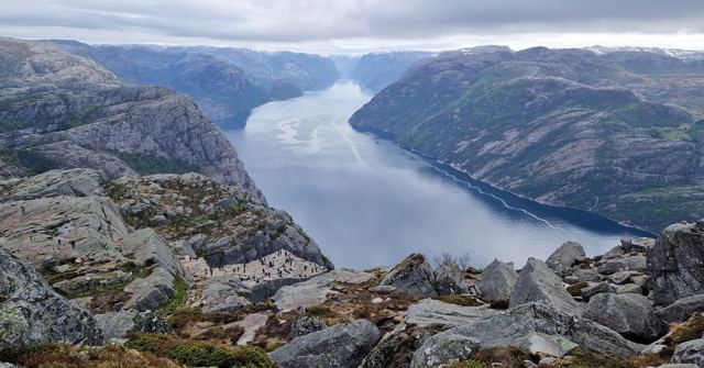 A long fjord winding between steep rocky mountains seen from a boulder-strewn summit.