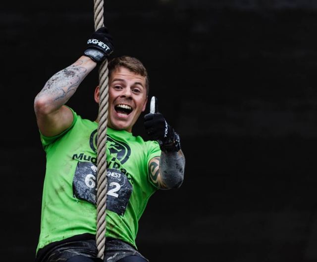 A man in a green race top climbing a rope and grinning during an obstacle race.