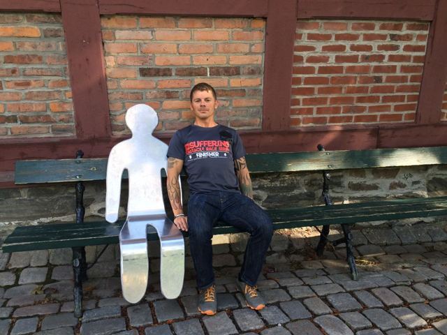 A man sitting on a bench beside a white figure-shaped cut-out against a brick wall.