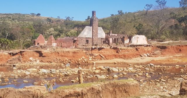 Ruined brick buildings beside a wide red stony riverbed with green hills behind.