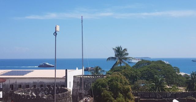 A rooftop view over buildings and palm trees to a turquoise sea with boats.