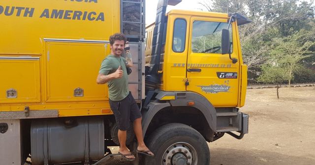 A man giving a thumbs up while standing on the step of a yellow overland truck.