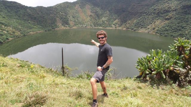 A man pointing towards a crater lake surrounded by green hills.