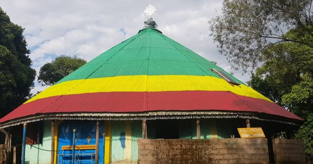 A round building with a green, yellow and red painted roof surrounded by trees.