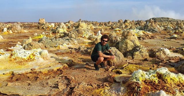 A man crouching among yellow and orange mineral formations under a blue sky.