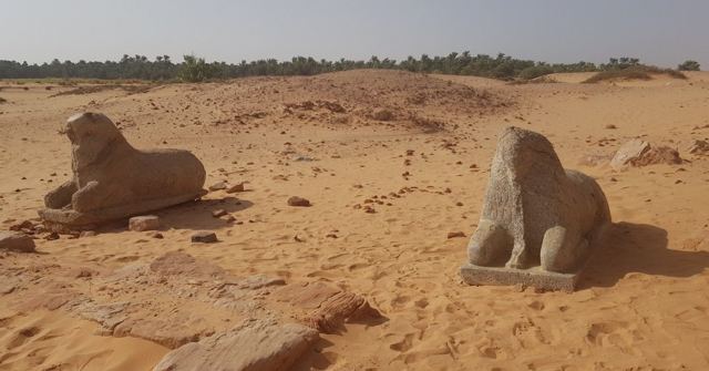 Two weathered stone statues of crouching animals on sand with palms in the distance.