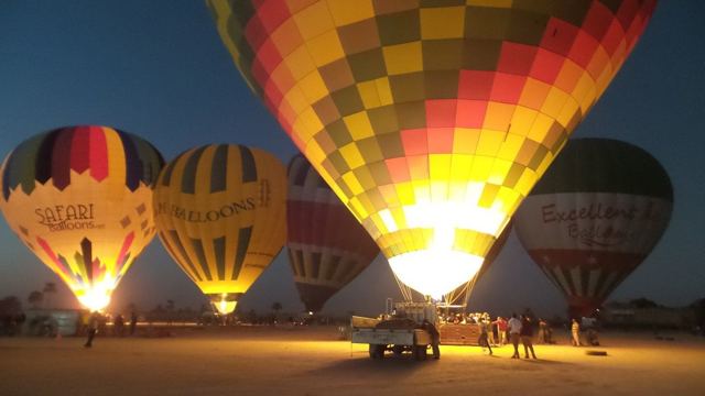 Several colourful hot air balloons being inflated and lit by burners on the ground before dawn.