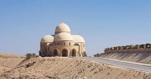 A domed white building on a rocky desert hill beside a road under blue sky.