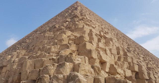 Looking up the stepped limestone blocks of a pyramid towards its peak against blue sky.
