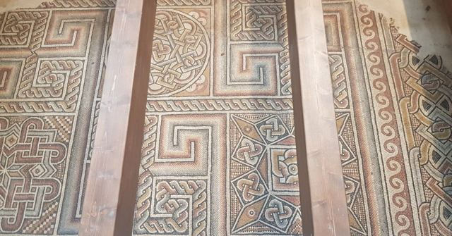Ancient geometric mosaic floor seen through gaps in wooden floorboards.