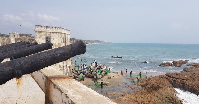 Black iron cannons on a white fort wall overlooking rocks, boats and the sea.