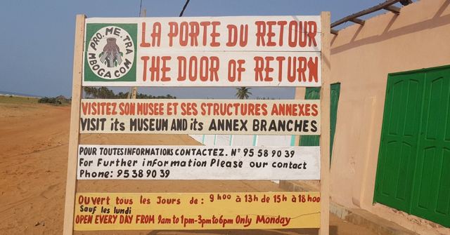 A sign reading La Porte du Retour, The Door of Return, with visiting information, by a green door.
