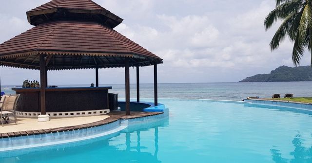A turquoise infinity pool with a thatched bar overlooking the sea and a distant island.