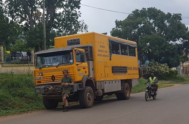 A yellow overland truck parked on a tree-lined road in Cameroon, a man beside it and a motorbike passing.