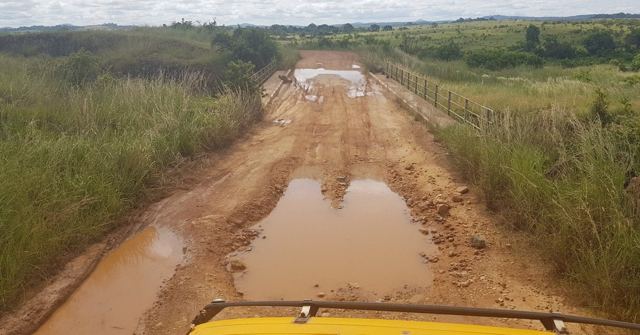 A muddy red dirt road full of puddles seen from the front of a yellow truck.