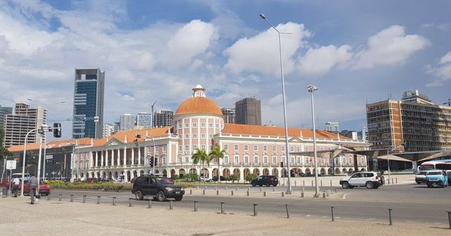 A pink and white colonial-style bank building with a red dome in front of modern skyscrapers in Luanda.