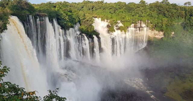 A wide waterfall pouring over a cliff into mist surrounded by green forest.