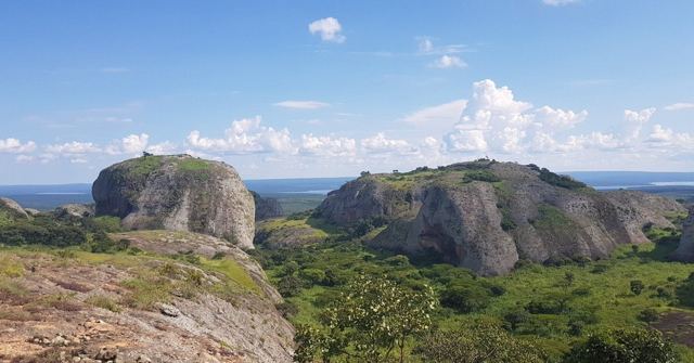 Huge rounded rock formations rising above green bushland under a blue sky.