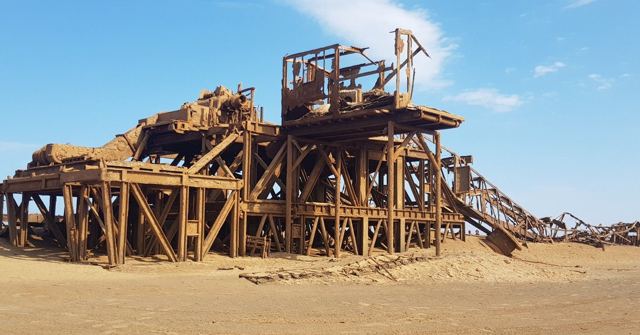 A rusted abandoned metal structure standing on sand under a blue sky.