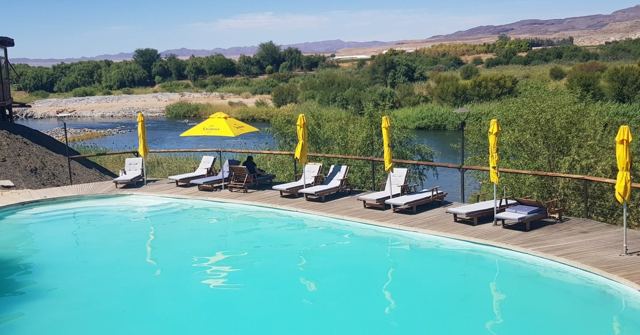 A bright blue swimming pool with sun loungers above a green riverbank and hills.