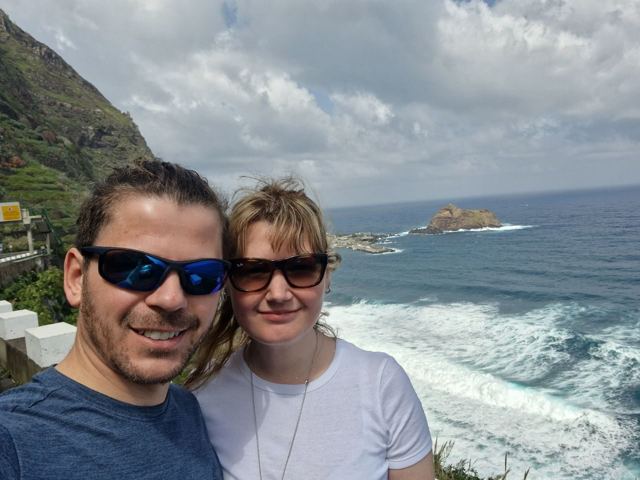 A man and a woman in sunglasses taking a selfie above crashing waves and sea stacks.