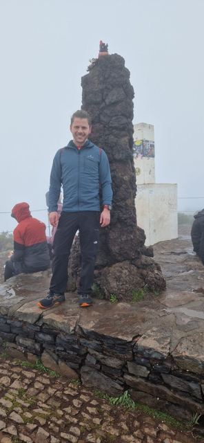 A man standing beside a stone summit pillar in the mist.