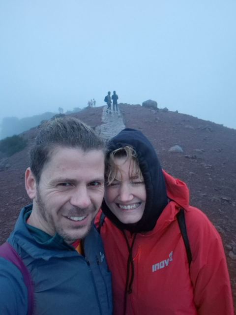 A smiling man and a woman in hoods taking a selfie on a red mountain path in the mist.