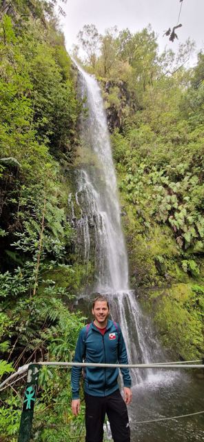 A man standing at the bottom of a tall waterfall in a lush green gorge.