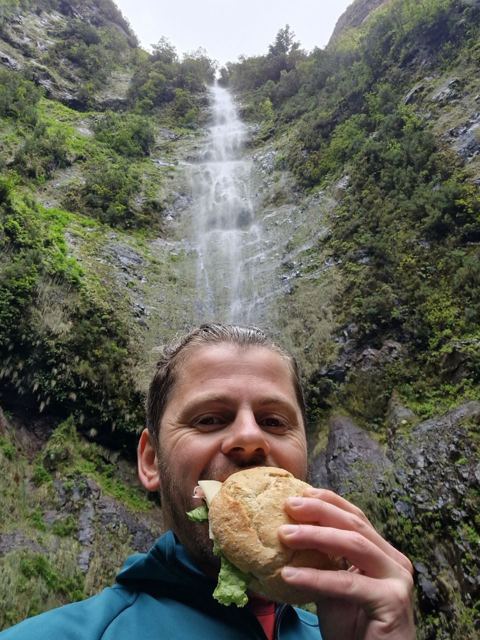 A man biting into a sandwich in front of a waterfall in green forest.