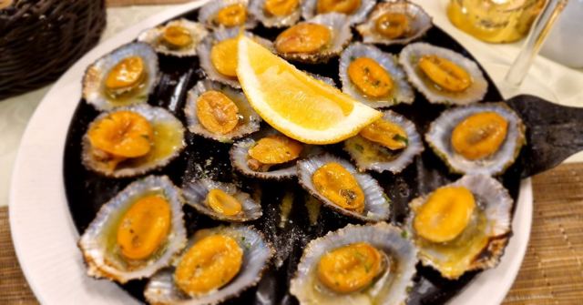 A dish of grilled limpets in their shells topped with butter and a lemon wedge.