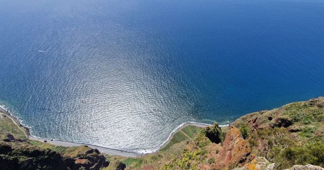 Looking down a sheer sea cliff to small coastal fields and a shimmering blue ocean.