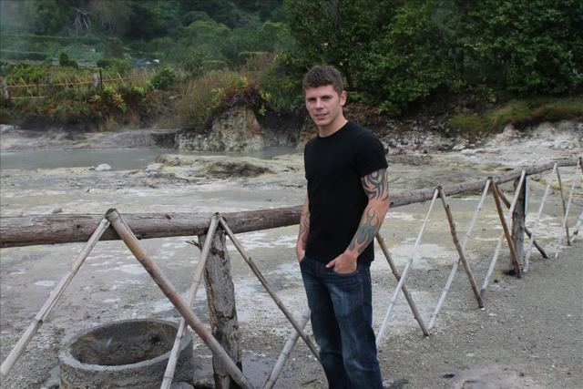 A man standing by a fence next to steaming volcanic hot springs.
