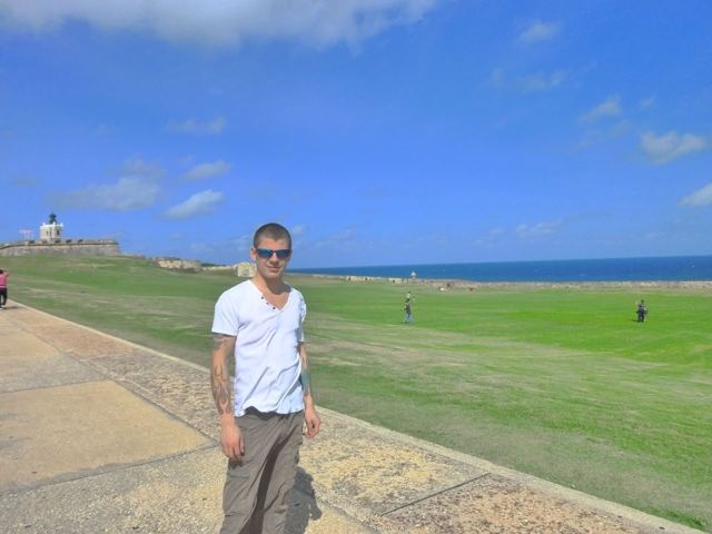 A man in a white T-shirt standing on the grassy lawn of El Morro fort beside the sea.
