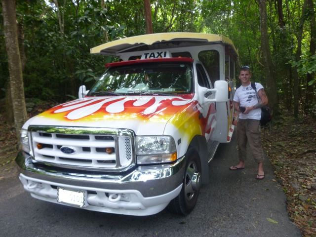 A man standing next to a flame-painted open-air safari taxi truck on a tropical road.