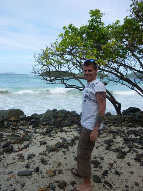 A man standing on a rocky beach under a windswept tree with the sea behind him.