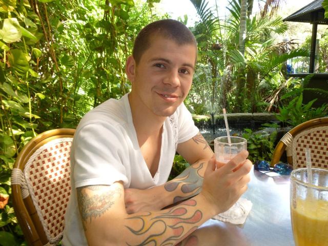 A smiling man with a tattooed arm holding a glass of juice at a café table surrounded by plants.