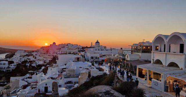 An orange sunset over the whitewashed buildings of Oia with crowds along the cliff.