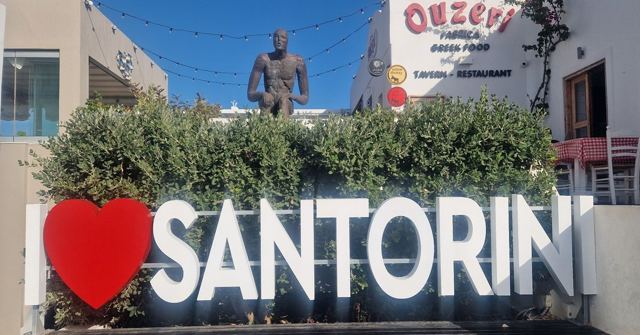 A large I love Santorini sign with a red heart, in front of a hedge and a statue.
