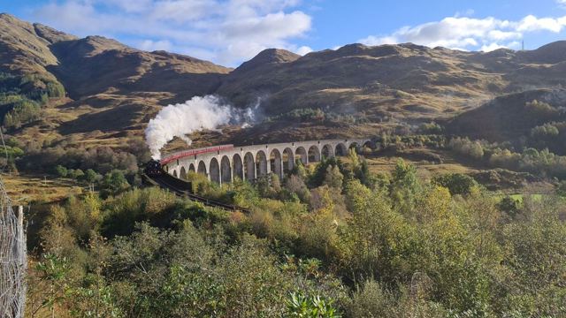 A steam train crossing a curved stone viaduct among autumn hills, trailing white smoke.