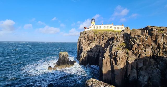 A white lighthouse on top of rugged sea cliffs above a deep blue, choppy sea.