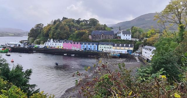 A row of brightly painted houses along a harbour, framed by trees and hills.