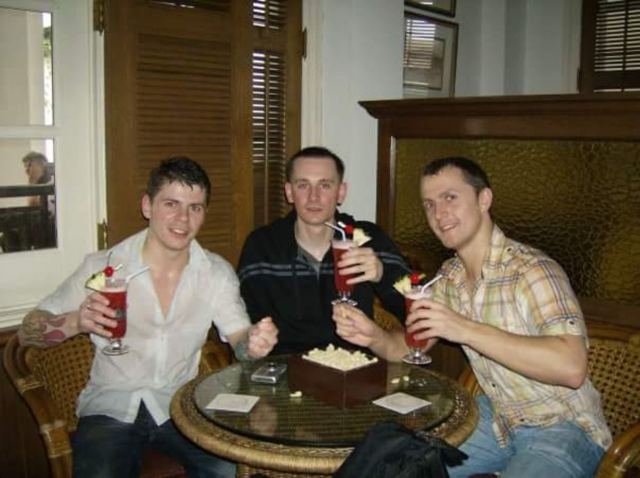 Three young men toasting with tall pink cocktails at a wooden table in a colonial-style bar in Singapore.