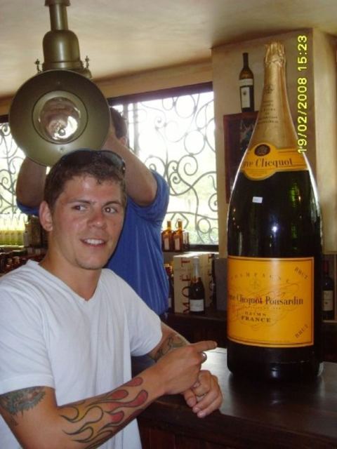 A smiling man in a white T-shirt standing beside a giant bottle of champagne in a wine shop.