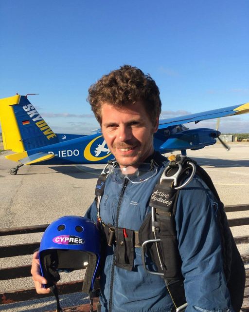 A smiling man in a skydiving jumpsuit and harness holding a helmet in front of a small plane.