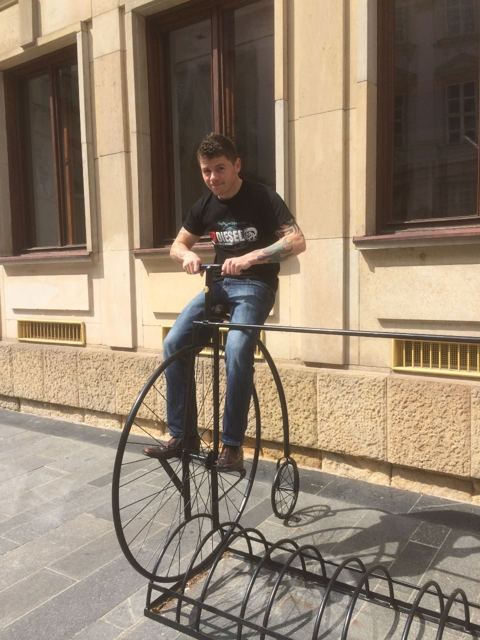A man sitting on an old-fashioned penny-farthing bicycle sculpture outside a building.