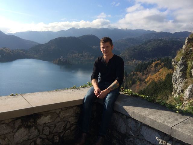 A man sitting on a stone wall above a lake surrounded by forested hills in Slovenia.