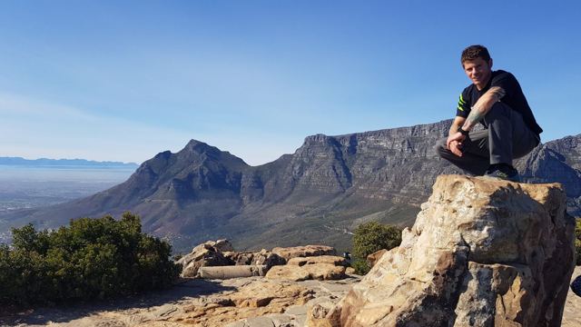 A man sitting on a rock at the summit of Lion's Head with Table Mountain behind.