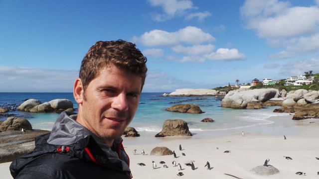 A smiling man taking a selfie with a white sand beach, boulders and penguins behind.